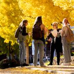 Instructor chats with students on U-N-C campus surrounded by bright yellow autumn leaves